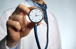 A doctor wears a stethoscope and holds the stethoscope bell up to the viewer. The bell has a clock face on it.