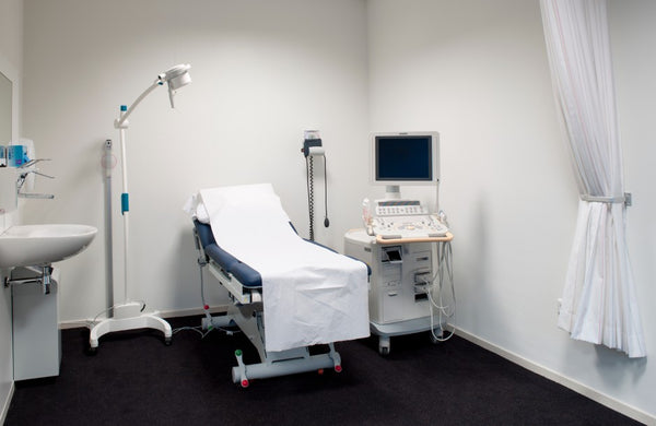 A white examination room with an electric exam table, ultrasound machine, blood pressure monitor, exam light, and sink.