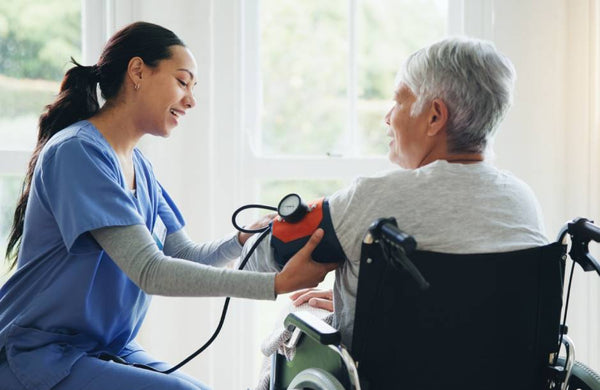 In a room with large windows, a healthcare worker places a blood pressure cuff on a senior patient sitting in a wheelchair.