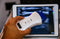 A hand holds a portable ultrasound probe. A portable ultrasound monitor displays a diagnostic image in the background.