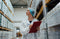 A woman wearing a lab coat, a hair net, and glasses walks through a medical supply warehouse with a clipboard.