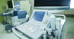 An ultrasound machine is set up in a room in a medical facility. Behind the machine is a desk with several screens.